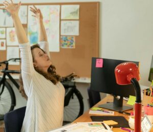 Lady middle aged stretching arms overhead while sitting at a computer desk. 