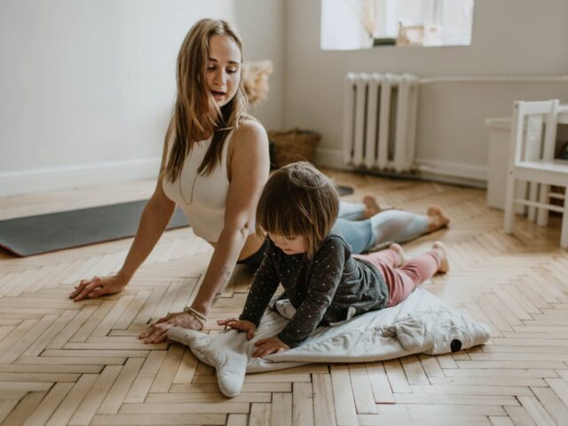 Lady with long hair in white singlet doign yoga exercise on floor with small child at home.