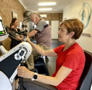 Lady with short hair in red shirt using arm cycle in gym group exercise class. 